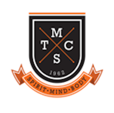 A black shield emblem with the letters T, M, C, and S separated by crossed lines, 1962 at the bottom, and an orange ribbon below reading SPIRIT • MIND • BODY.