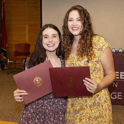 Two young women smiling and holding certificates stand side by side in a room with a wooden wall and a table in the background. Both are dressed in floral-patterned dresses.