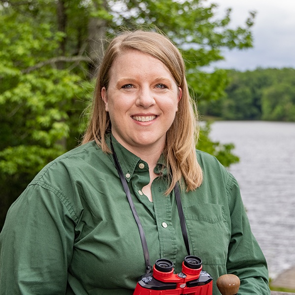 A woman with blonde hair wearing a green button-down shirt stands outdoors near a lake, holding red binoculars around her neck. Lush green trees and water are in the background.