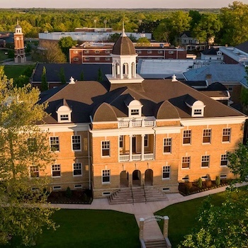 Aerial view of a large brick building with a central cupola, arched entryway, and symmetrical windows, surrounded by green trees and lawns, with additional campus buildings in the background.