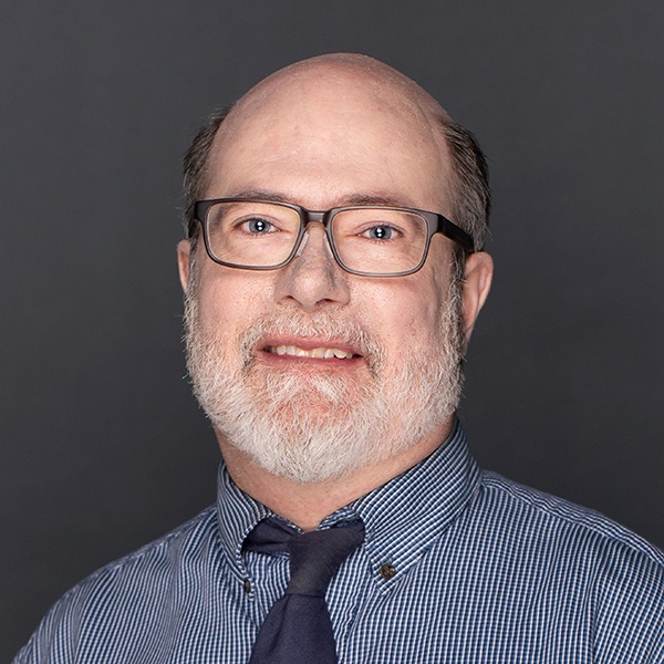 A middle-aged man with glasses, a gray beard, and short hair smiles at the camera. He is wearing a blue checked shirt, a navy tie, and is posed against a plain dark gray background.