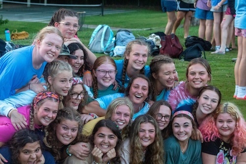 A group of smiling young women pose closely together outdoors, some kneeling and some standing. They appear happy and are gathered on a grassy area with backpacks in the background.