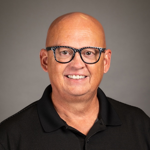 A smiling bald man wearing clear glasses and a striped polo shirt with a blue collar, posing in front of a plain gray background.