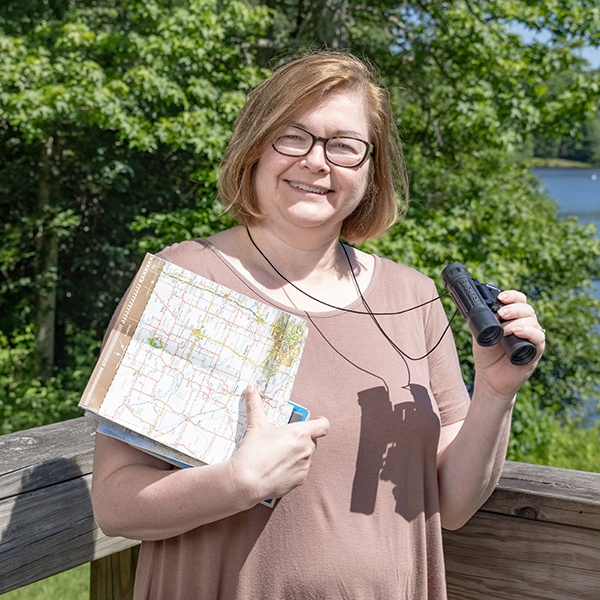 A woman with glasses stands outdoors by a wooden railing, holding a map and a book in one arm and binoculars in the other. Trees and a body of water are visible in the background on a sunny day.
