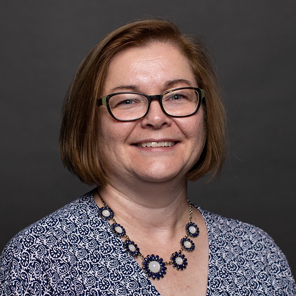 A woman with short brown hair and glasses smiles at the camera. She is wearing a blue patterned blouse and a necklace with circular blue and white designs. The background is plain and dark.
