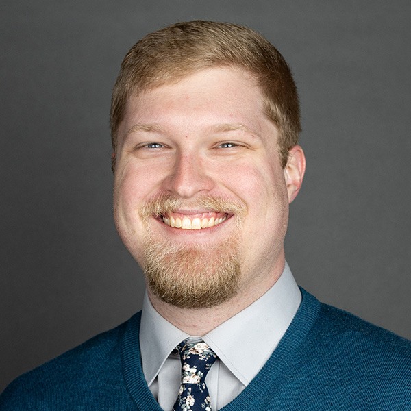 A man with short light brown hair, a beard, and mustache smiles at the camera. He is wearing a teal sweater over a light gray collared shirt and a dark floral tie. The background is plain and gray.