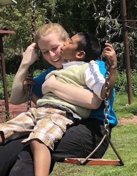 A smiling woman sitting on a swing holds a young boy in her lap. The boy is giving her a kiss on the cheek. They are outdoors on a sunny day, surrounded by green grass and trees.
