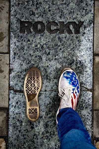 A person wearing jeans and a sneaker with an American flag design stands next to a shoe print on a stone slab engraved with the word “ROCKY.” The scene mimics the iconic Rocky movie footprint.