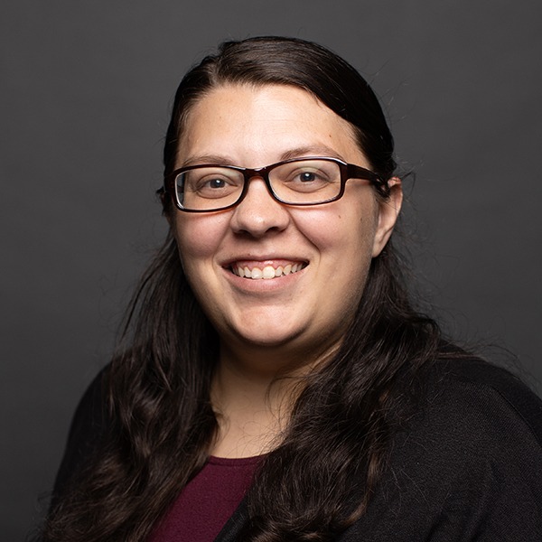 A woman with long dark hair and glasses smiles at the camera. She is wearing a dark top and a black cardigan, with a plain dark background behind her.