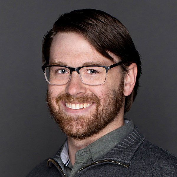 A man with short brown hair, a beard, and glasses smiles at the camera. He is wearing a dark zip-up sweater over a collared shirt with a plain dark background.