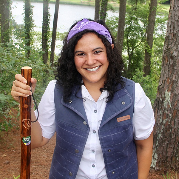 A smiling woman with curly hair, wearing a purple bandana, white shirt, and blue vest, stands in a forest holding a wooden walking stick, with a lake visible in the background.