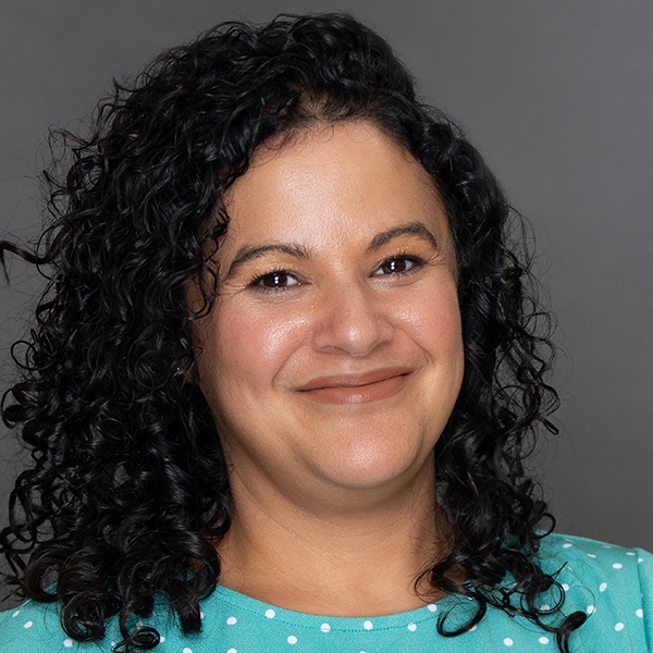 A woman with curly dark hair smiles at the camera. She is wearing a light turquoise top with white polka dots and is posed in front of a plain gray background.