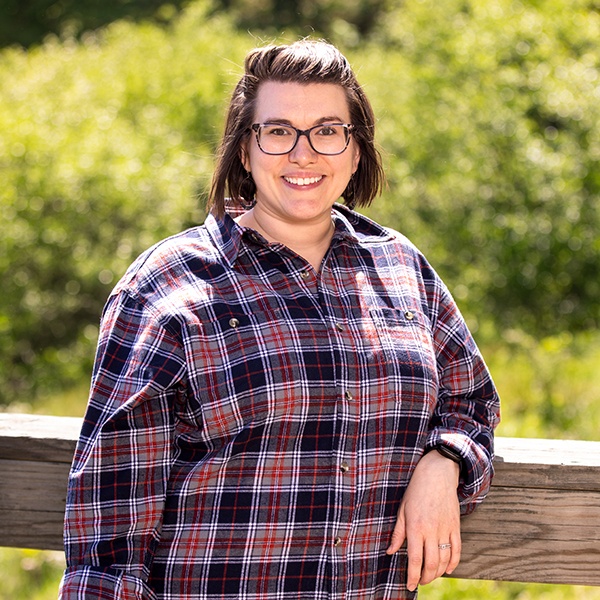 A person wearing glasses and a red and blue plaid shirt stands outdoors, smiling, with one hand resting on a wooden fence. Lush green foliage is visible in the background.