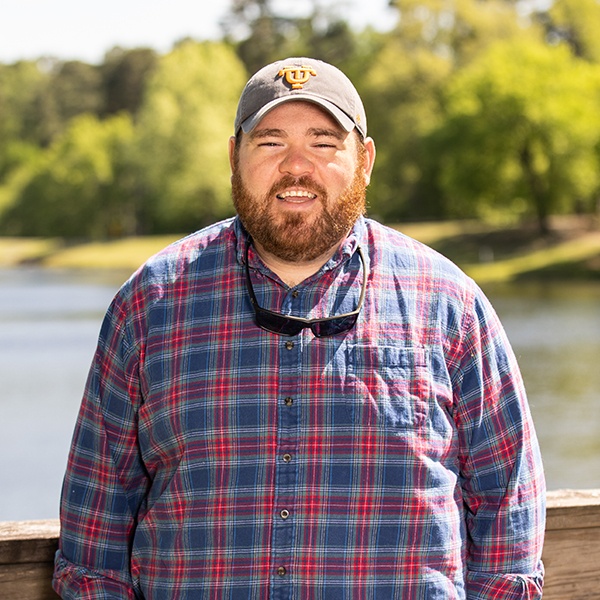 A bearded man in a plaid shirt and baseball cap stands outdoors on a wooden bridge with a lake and green trees in the background on a sunny day.