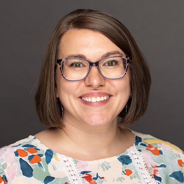 A woman with straight brown hair, wearing glasses and a floral patterned blouse, smiles at the camera against a plain dark background.