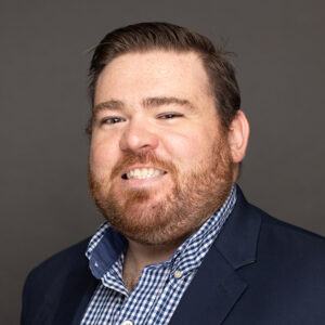 A man with short brown hair and a beard smiles at the camera. He is wearing a dark blazer over a blue and white checkered shirt, in front of a plain gray background.