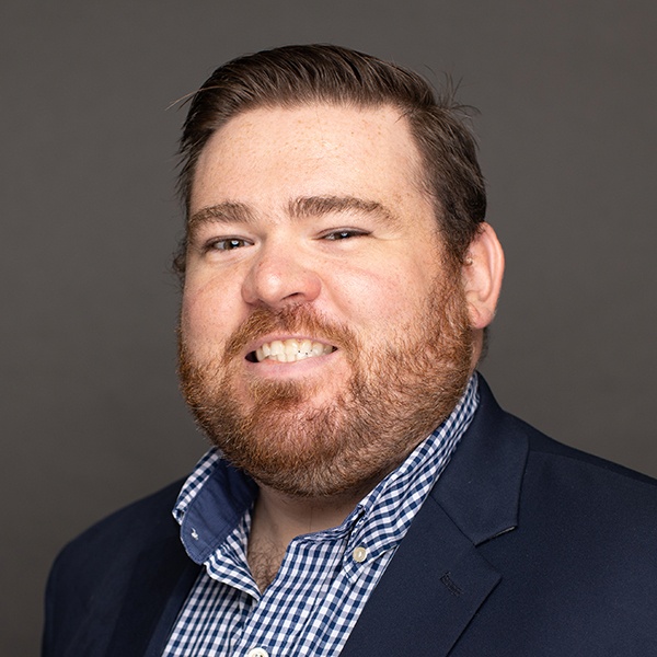 A man with short brown hair and a beard smiles at the camera. He is wearing a dark blazer over a blue and white checkered shirt, in front of a plain gray background.