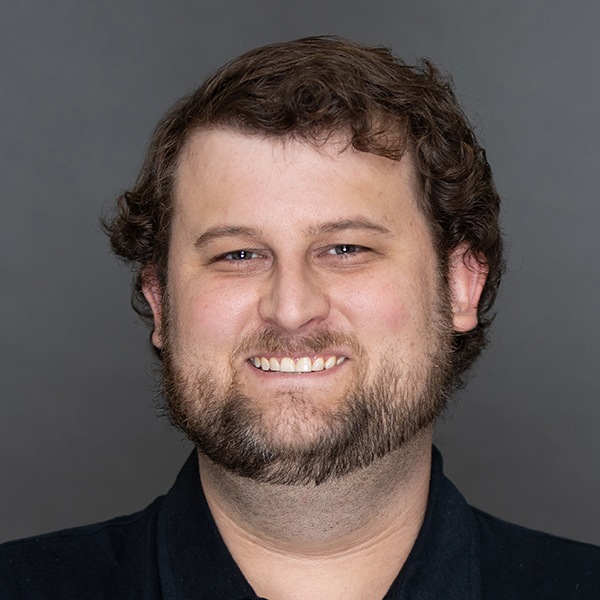A man with curly brown hair, a full beard, and a mustache is smiling at the camera. He is wearing a dark collared shirt and is posed in front of a plain gray background.