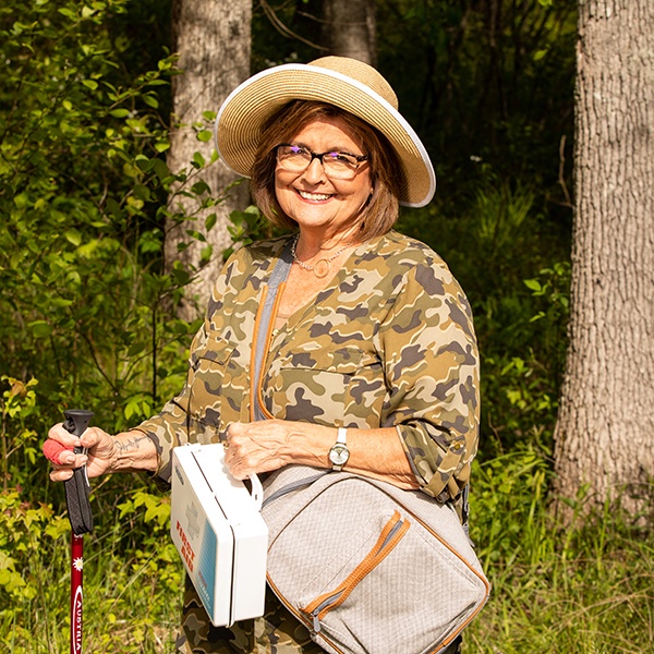 Smiling older woman wearing a camouflage shirt, hat, and glasses stands outdoors in a sunny forest, holding a walking stick and carrying a bag and a small cooler.