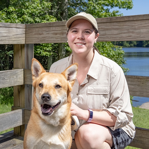 A woman wearing a beige shirt and cap sits on a wooden deck by a lake, smiling at the camera while holding a tan and white dog. Trees and blue water are visible in the background on a sunny day.