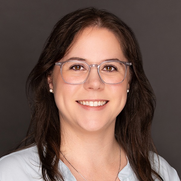 A woman with long brown hair, wearing clear glasses, pearl earrings, and a light blue blouse, smiles at the camera against a plain dark background.