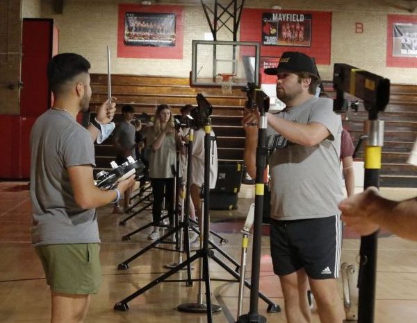 Several people stand in two rows facing each other in a gym, each holding fencing equipment and wearing casual clothes. A basketball hoop and wooden bleachers are visible in the background.