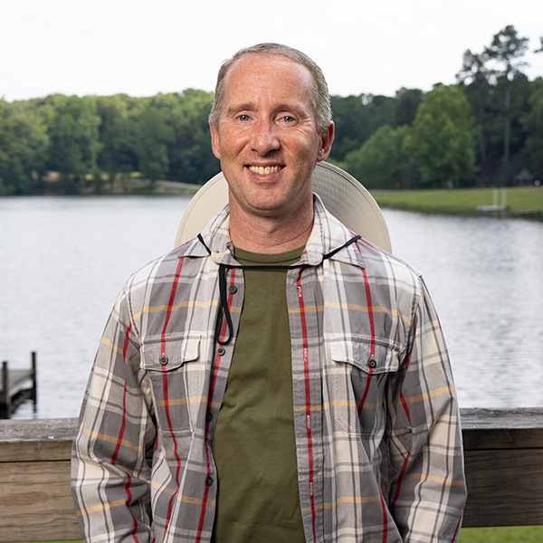 A smiling man in a plaid shirt and green t-shirt stands on a wooden deck by a lake, with trees and a dock visible in the background on a bright day.