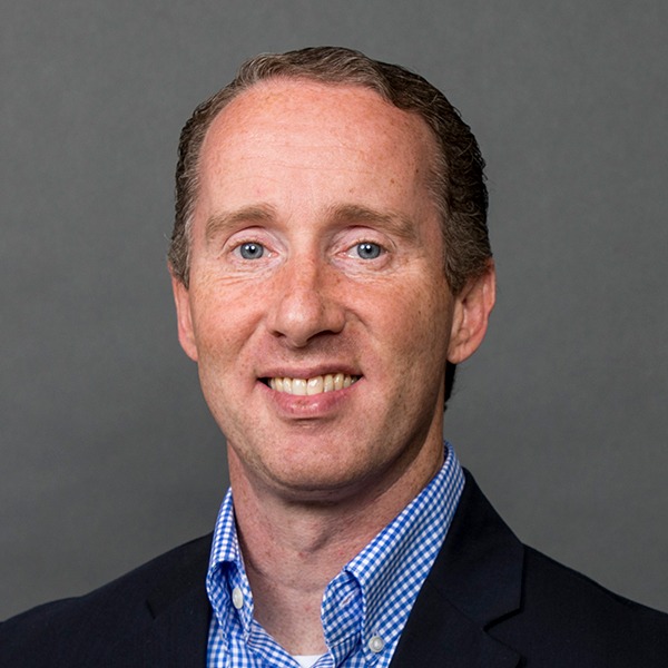 A man with short brown hair wearing a dark suit jacket, blue and white checkered dress shirt, and no tie, smiling in front of a plain gray background.