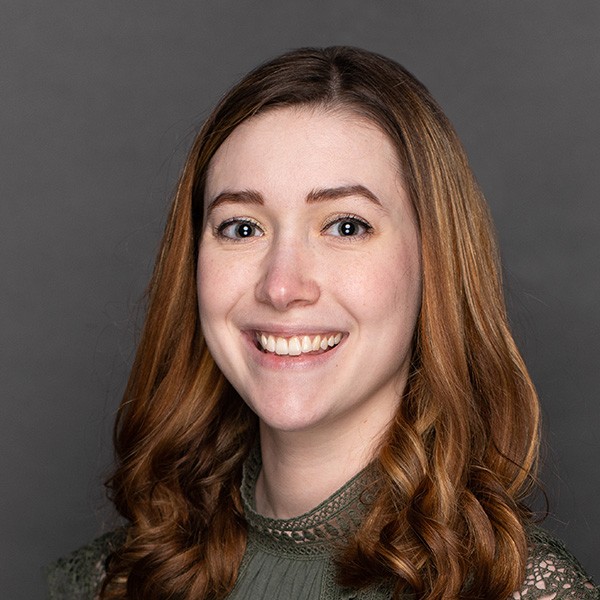 A young woman with long, wavy auburn hair smiles at the camera. She is wearing a green, high-necked top and is posed against a plain gray background.