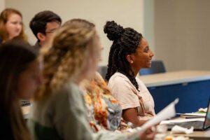 Students sit in a classroom, listening attentively. A woman with braided hair smiles, facing forward. Desks, papers, and a laptop are visible in the scene. The image captures an engaged learning environment.