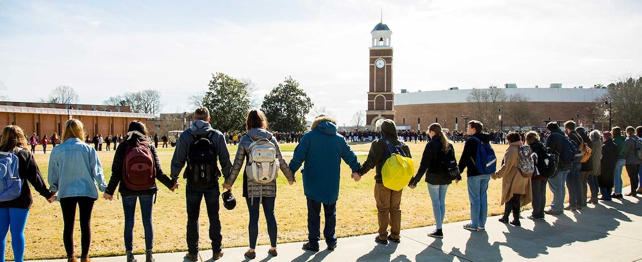 A diverse group of people stands outdoors in a large circle, holding hands on a grassy field near a clock tower and brick buildings, under a clear sky.