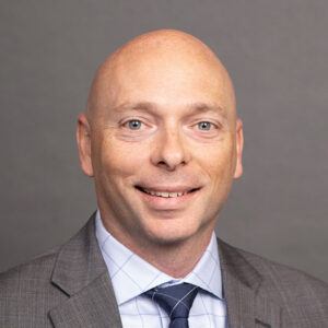A bald man in a gray suit, white shirt, and striped tie smiles at the camera against a plain gray background.