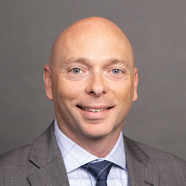 A bald man in a gray suit, white shirt, and striped tie smiles at the camera against a plain gray background.