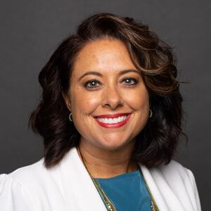 A woman with shoulder-length wavy brown hair and highlights, wearing a white blazer over a teal top, smiles at the camera against a plain dark background.
