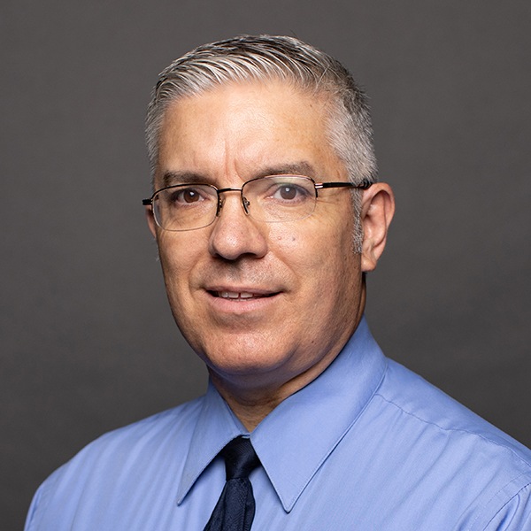 A middle-aged man with short gray hair, glasses, and a clean-shaven face wears a light blue collared shirt and dark tie, posing in front of a plain gray background.