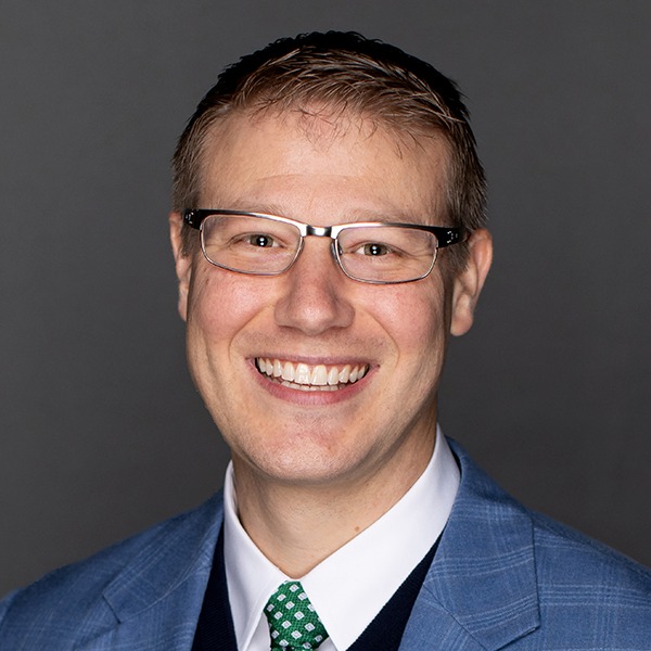 A man wearing glasses, a blue suit jacket, a white shirt, and a green patterned tie, smiling in front of a dark gray background.