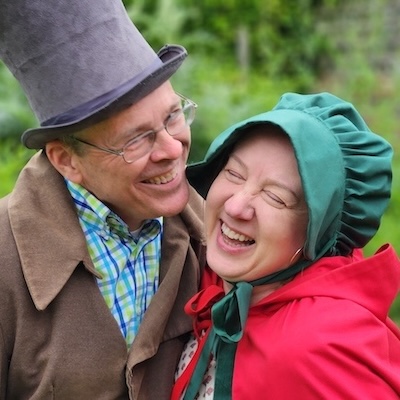 A man and a woman dressed in historical ‘Romantic England’ style costumes sharing a joyful moment, outdoors. The man wears a tall hat and a brown coat; the woman wears a bonnet and a red cloak. Both are smiling and laughing.