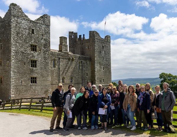 A group of people pose for a photo in front of a large, historic stone castle under a partly cloudy sky, with green hills and a wooden fence in the background.