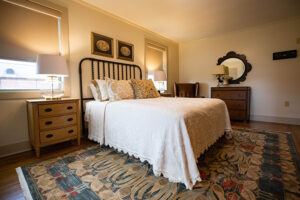 A cozy bedroom features a double bed with white bedding, a wooden headboard, two bedside tables with lamps, a dresser with a round mirror, floral artwork, and a patterned area rug. Warm light fills the room.