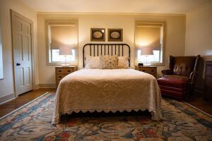 A cozy bedroom with a double bed, cream bedding, two bedside tables with lamps, framed art above the headboard, an armchair, and a colorful rug on a wooden floor.