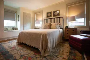 A cozy bedroom with a metal-framed bed, cream bedding, two bedside tables with lamps, a cushioned window seat, a patterned rug, and a red leather armchair and ottoman. Natural light enters through the windows.