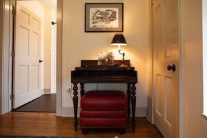 A small wooden desk with a lamp, decor items, and framed art above it sits against a wall. Two red cushioned stools are placed underneath. Doors stand open on either side, letting in natural light.