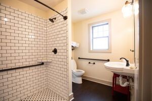 Accessible bathroom with a walk-in shower featuring grab bars and white subway tiles, a toilet with support bars, a window, and a pedestal sink with a wall-mounted light above it.