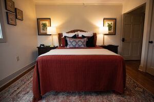 A cozy bedroom with a large bed covered in a rust-colored blanket, decorative pillows, two bedside tables with lamps, framed art on the wall, and a patterned rug on the wooden floor.