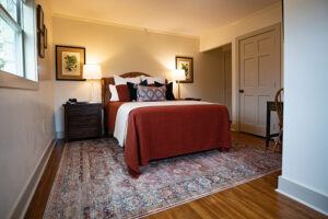A cozy bedroom with a double bed featuring red and white bedding, two bedside tables with lamps, framed artwork on the wall, a patterned rug, and wooden floors. Natural light enters from a window on the left.
