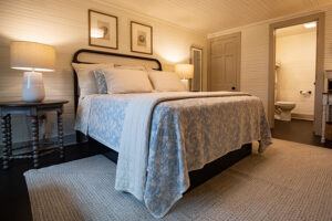 A cozy bedroom with a double bed covered in a light patterned quilt, two bedside tables with lamps, framed artwork above the bed, and an open door leading to a bathroom.