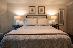 A neatly made bed with a patterned comforter and multiple pillows is centered between two nightstands, each with a lit lamp. Two framed pictures hang above the headboard on a white paneled wall.