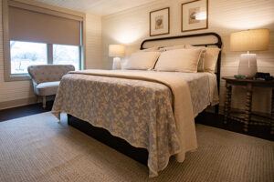 A cozy bedroom with a neatly made bed, two bedside tables with lamps, framed art above the headboard, a patterned cream bedspread, and a cushioned chair by a window with blinds.