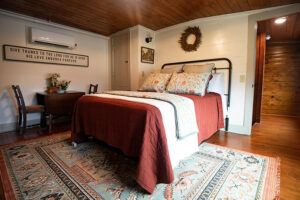 Cozy bedroom with a metal-frame bed, patterned pillows, and a rust-colored blanket. There’s a large rug, wooden ceiling, small table and chairs, wall decor, and warm lighting creating an inviting atmosphere.