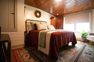 A cozy bedroom with a wooden ceiling, a bed with red and cream bedding, decorative pillows, a rug on the floor, a window with a shade, a green plant, and a wreath on the wall above the bed.